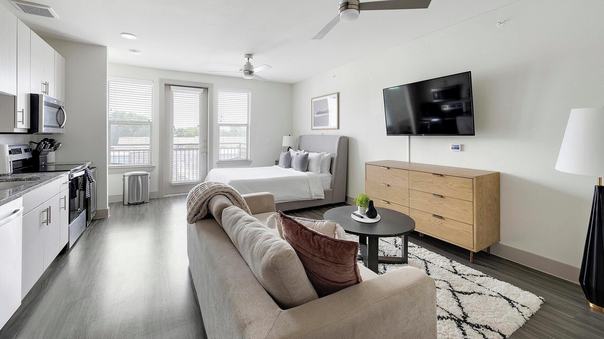 A modern hotel room with a kitchenette, a bed, a sofa, a TV, and a wooden cabinet.
