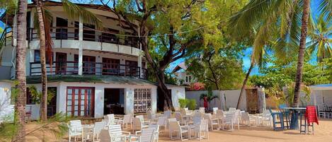 A beachfront property with a two-story building, outdoor seating area with white chairs, and palm trees.