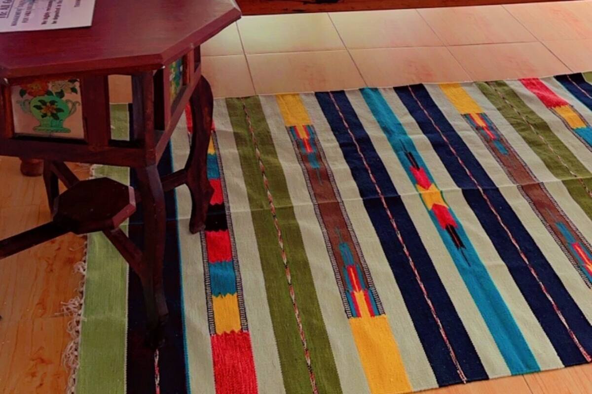 A bedroom with a bed, a wooden table with a vase of flowers, and a colorful striped rug.
