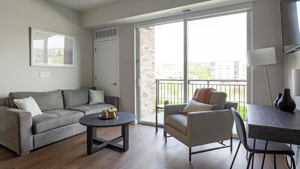A living room with a grey sofa, a round coffee table, a grey armchair, and a small dining table with chairs. There is a large window with a view of buildings and a balcony outside.