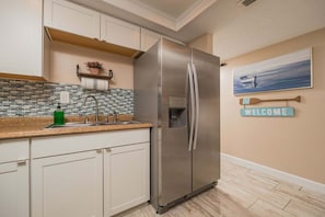 A kitchen with a stainless steel refrigerator, a sink with a faucet, a tiled backsplash, and a welcome sign on the wall. - Pelican Inlet D229- The Chubby Mermaid (St. Augustine)