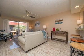A living room with a sofa, a coffee table, a dining table with stools, a ceiling fan, and a balcony with a view. - Pelican Inlet D229- The Chubby Mermaid (St. Augustine)