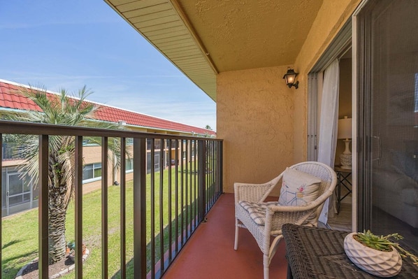 A balcony with a wicker chair, a small table with a plant, and a view of a building with a red roof. - Pelican Inlet D229- The Chubby Mermaid (St. Augustine)