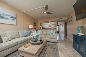 A living room with a sofa, coffee table, and a ceiling fan. There is a kitchen in the background. - Pelican Inlet D229- The Chubby Mermaid (St. Augustine)