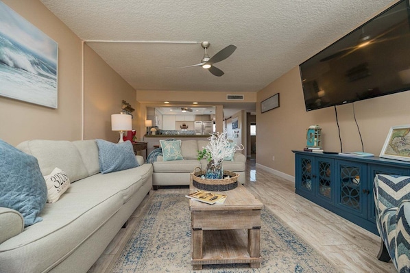 A living room with a sofa, coffee table, TV, and a view into the kitchen. - Pelican Inlet D229- The Chubby Mermaid (St. Augustine)