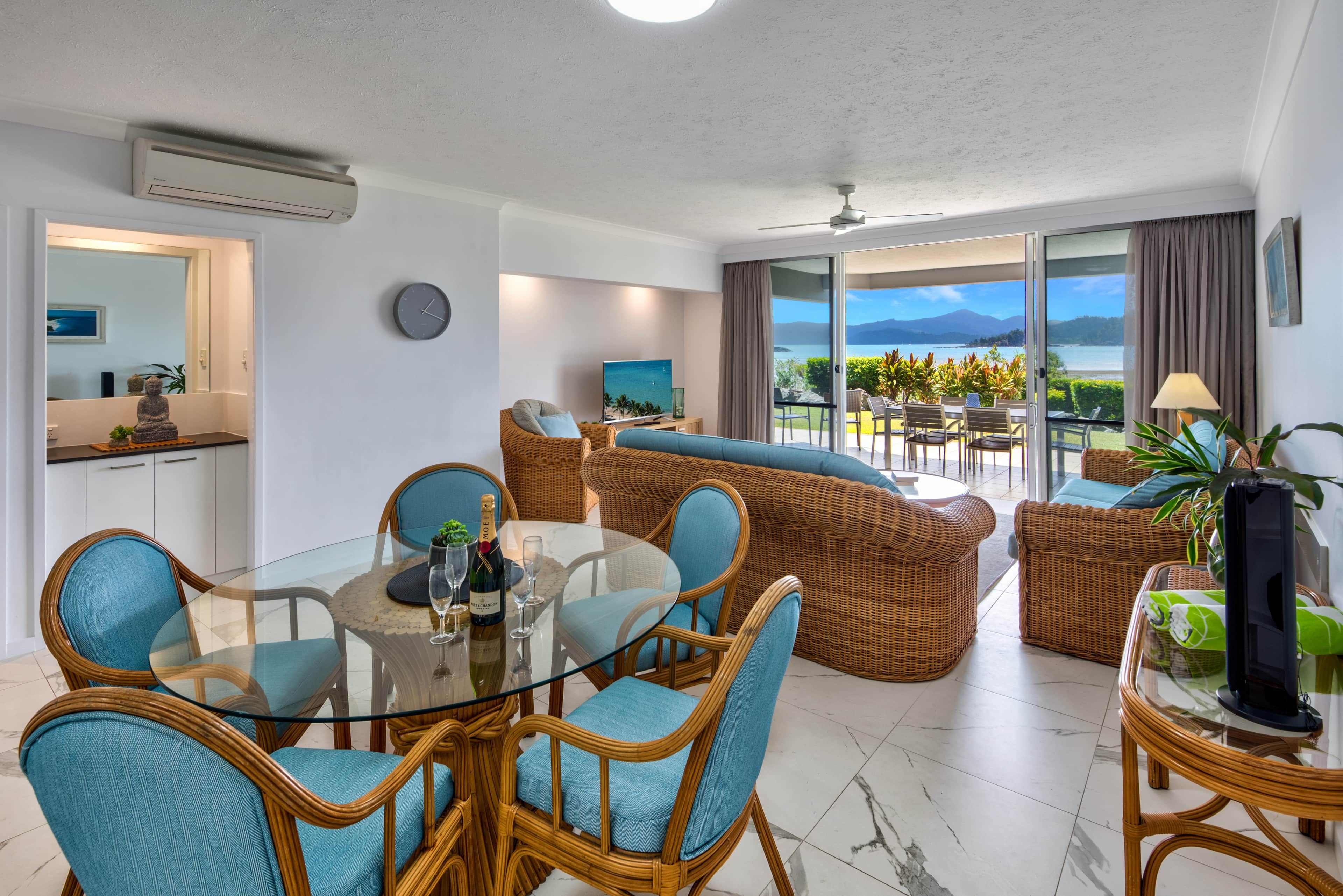 A modern living room with a glass dining table, wicker furniture, and a view of the sea.