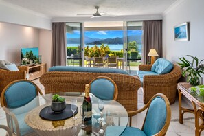 A modern living room with a glass table, wicker furniture, and a scenic view of a lake and mountains. - Frangipani Lodge F5, Frangipani F5 on Hamilton Island (Hamilton Island)