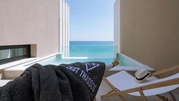 Un balcón con vista al océano, una toalla con el texto 'ZEST THASSOS' y un sombrero en una silla.