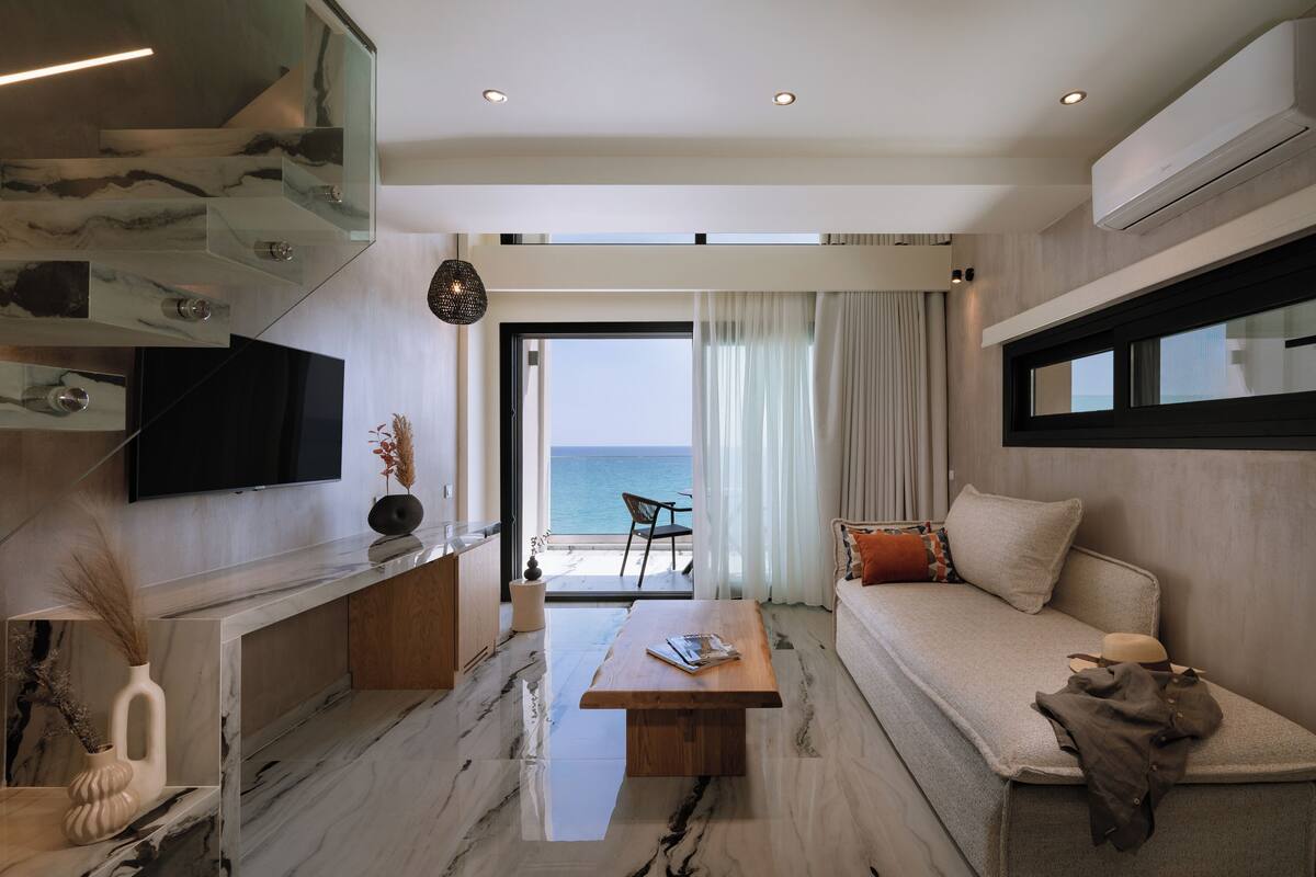 A modern living room with a marble floor, a flat-screen TV, a wooden coffee table, a sofa, and a view of the sea.