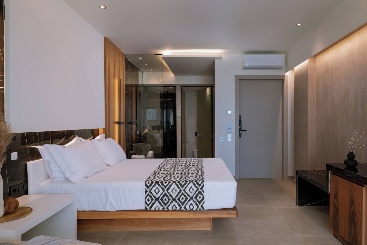 A modern hotel room with a large bed, a bedside table, a wardrobe, and a bathroom visible through a glass door.