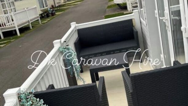 A balcony with two black wicker chairs, a white railing, and a hanging plant. - Luxury Caravan in Towyn North Wales (Abergele)