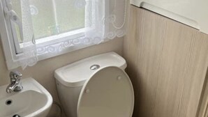 A bathroom with a toilet, sink, and window with a decorative curtain. - Luxury Caravan in Towyn North Wales (Abergele)