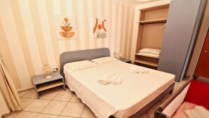 A neatly made bed with pillows and rolled towels, a bedside table with a lamp, a wardrobe, and two wall art pieces.