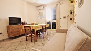 Een woonkamer met een bank, een eettafel met stoelen, een televisie op een houten kast en een raam met gordijnen.