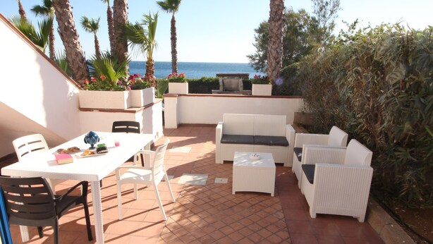 Un patio con mobiliario de mimbre blanco, una mesa con un jarrón azul y vista al mar.