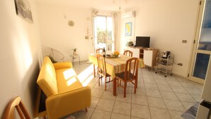 Un comedor luminoso con un sofá amarillo, una mesa de comedor de madera y sillas. Hay un televisor sobre un mueble bajo y una pequeña mesita con una planta.
