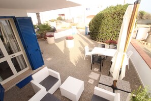 A patio area with white wicker furniture, a table, and chairs. There is a blue door and a white wall with a window.
