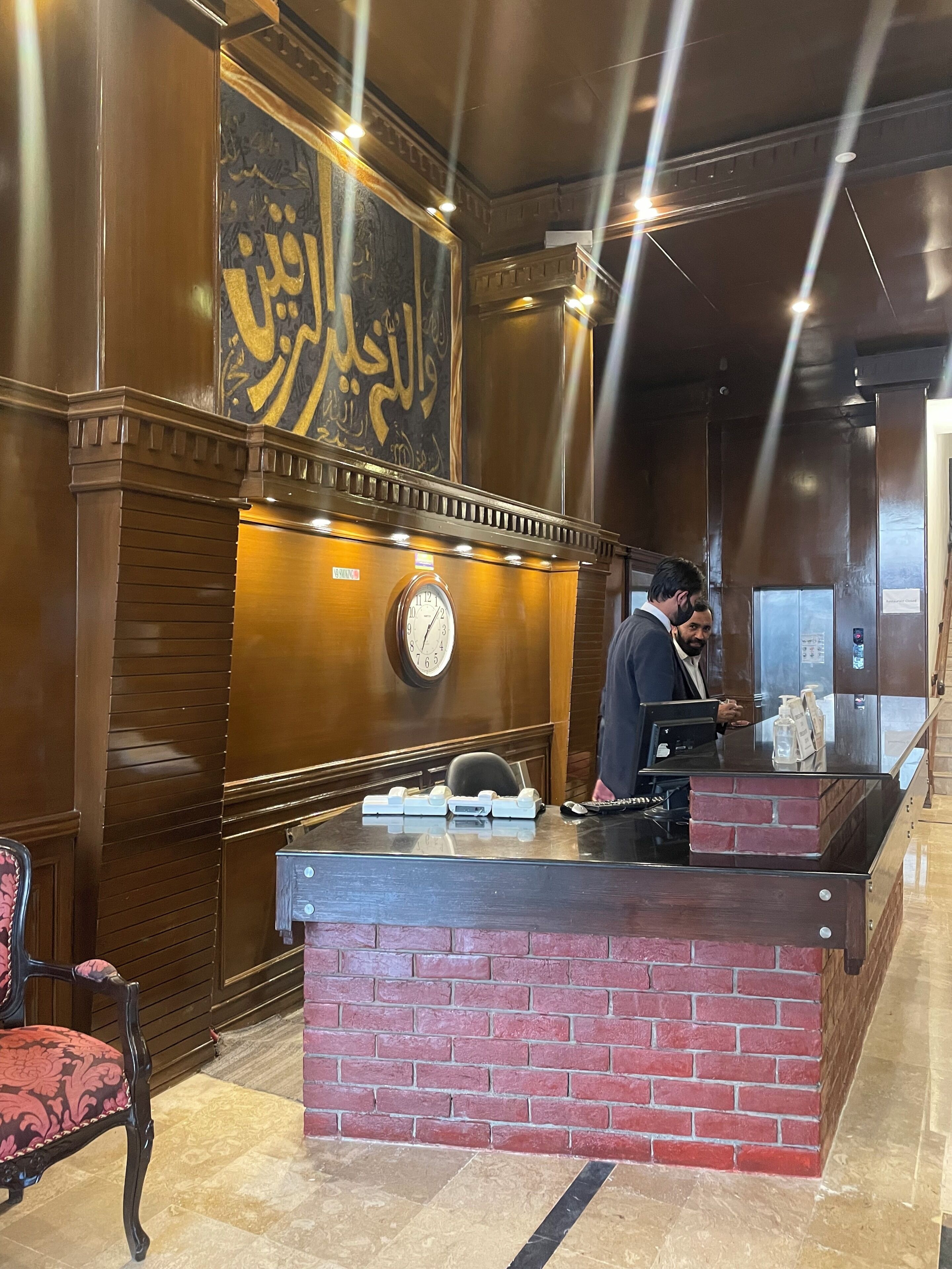 Een receptiegedeelte met een houten receptiebalie, een klok en een muur met Arabische kalligrafie.