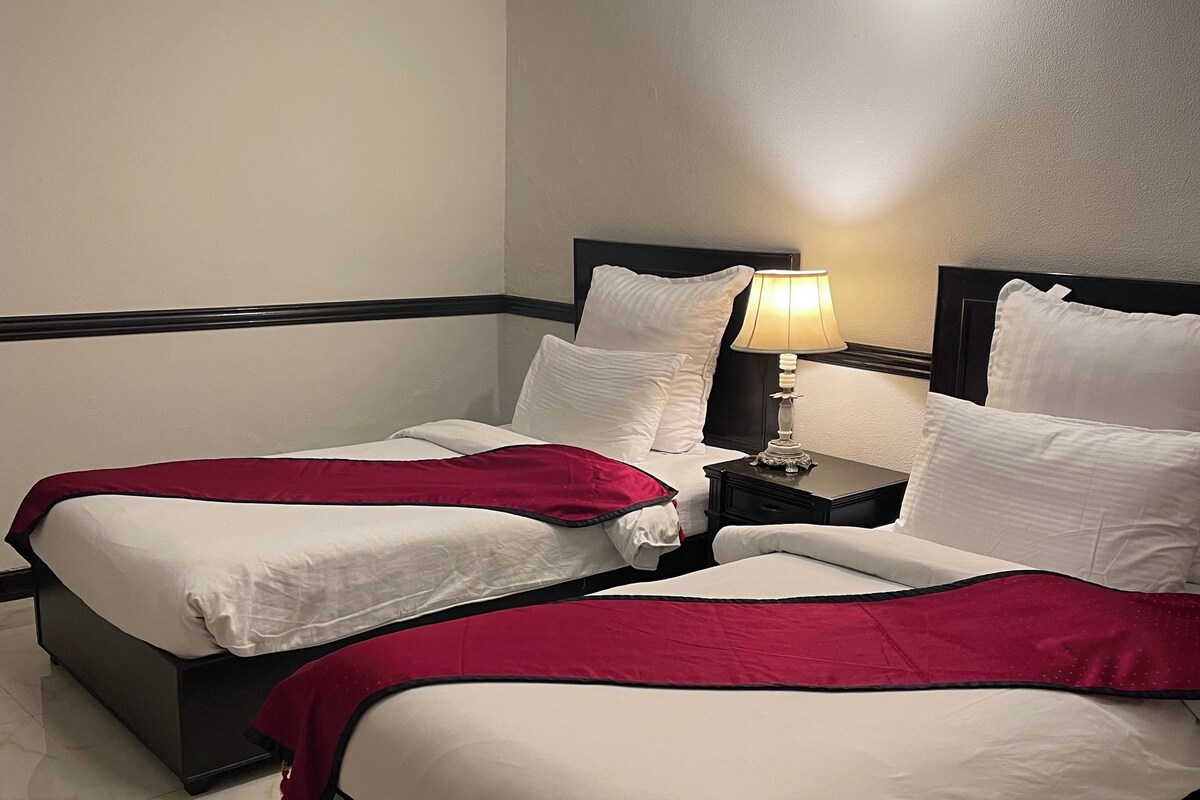 Habitación de hotel con dos camas, una mesita de noche con lámpara e iluminación en el techo.