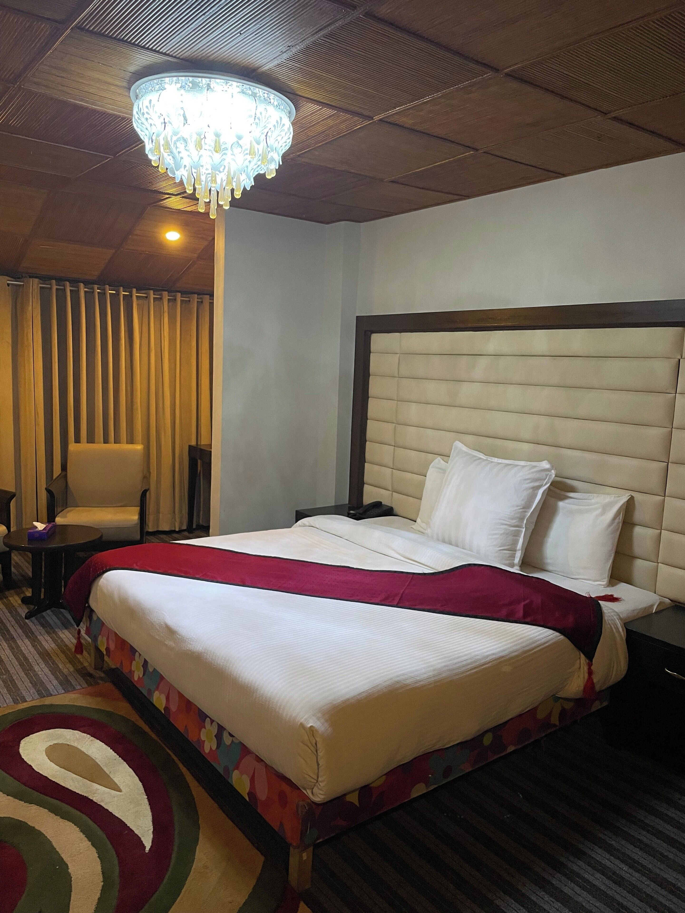 Ein Hotelzimmer mit einem großen Bett, einem Sessel, einem Nachttisch, einer Kristallleuchtre und einem gemusterten Teppich.