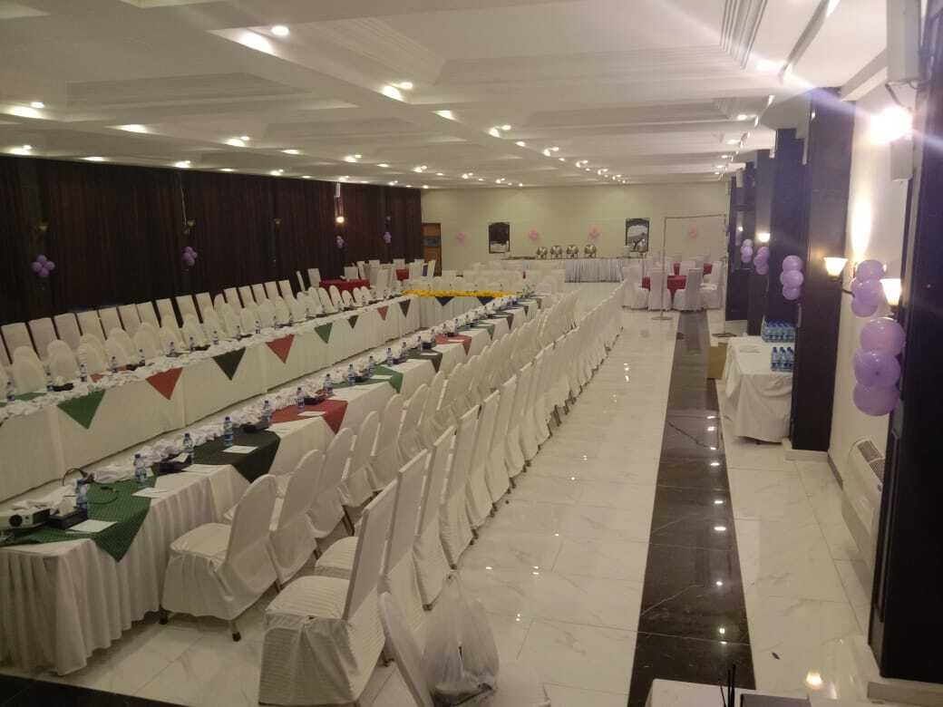 Un salón de banquetes con filas de mesas y sillas, un escenario y una mesa larga con mantel verde.
