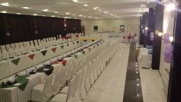 Un salón de banquetes con filas de mesas y sillas, un escenario y una mesa larga con mantel verde.