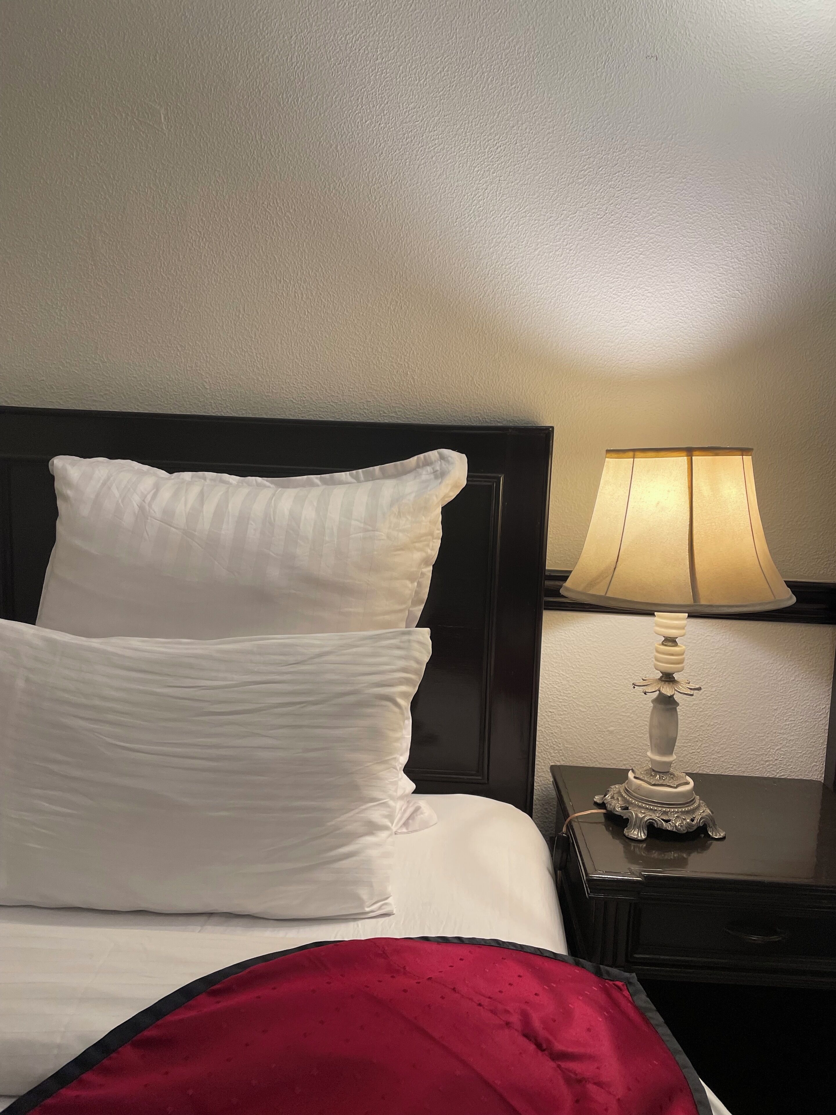 Una cama bien hecha con almohadas blancas y una manta roja, una mesita de noche negra con una lámpara, y una pared lisa de fondo.