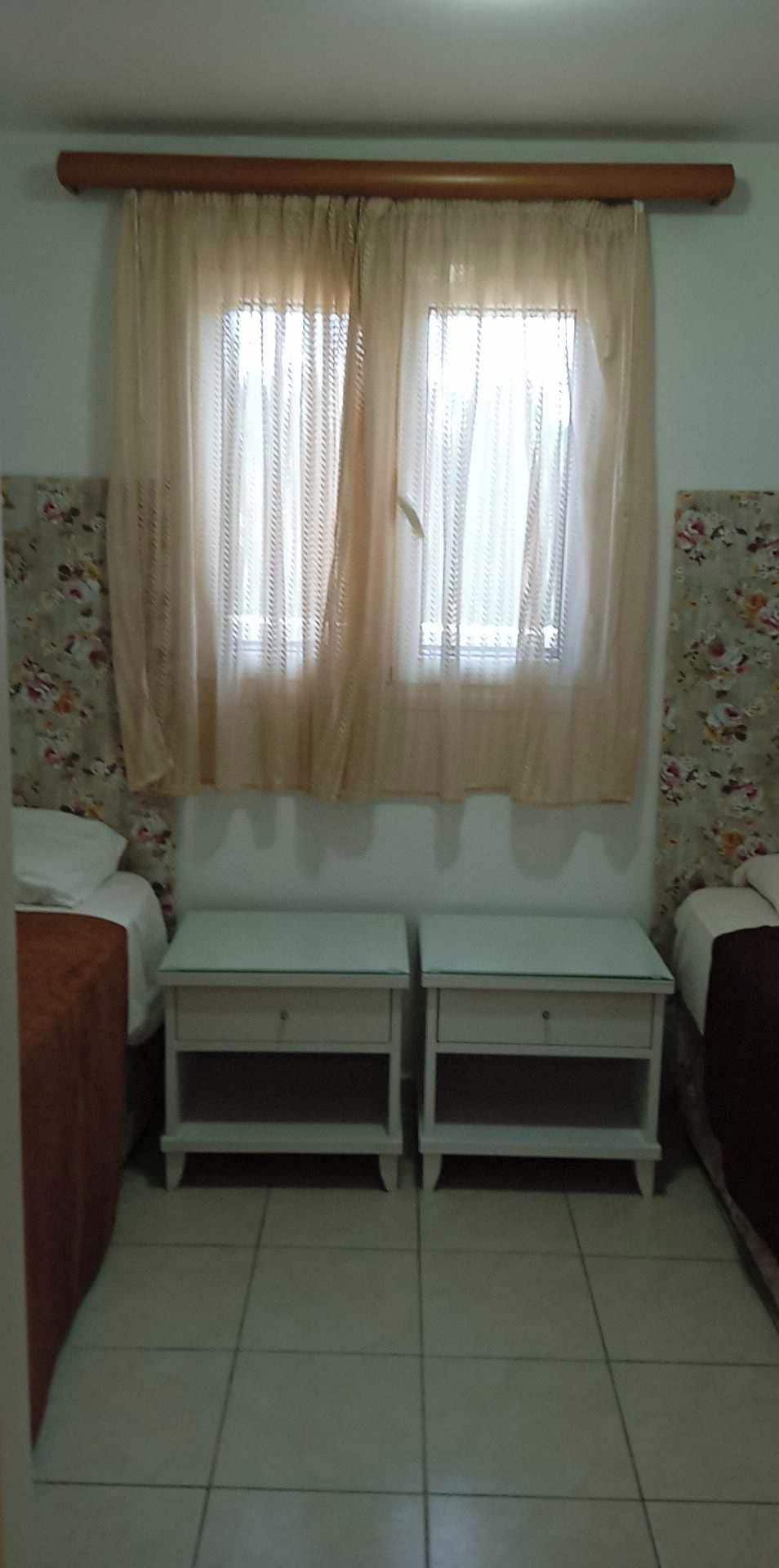 A room with two bedside tables, a window with curtains, and floral wallpaper.