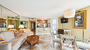 A modern living room with a sofa, coffee table, and glass dining table. There is a kitchen in the background with a TV mounted above the counter. - Includes Xplorie Activities and Linens. PYRAMID - 1BR Veranda condo w/outdoor pool. (Ocean City)