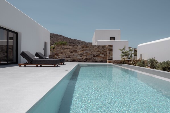 A modern pool area with a clear blue pool, lounge chairs, and a stone wall. - Ios Bliss (Ios)