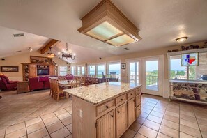 A spacious living area with a large island, wooden cabinets, a chandelier, and a view of the outdoors.