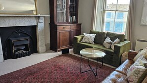 Un salon avec un canapé vert, une petite table ronde, une cheminée et une grande fenêtre donnant sur des immeubles.