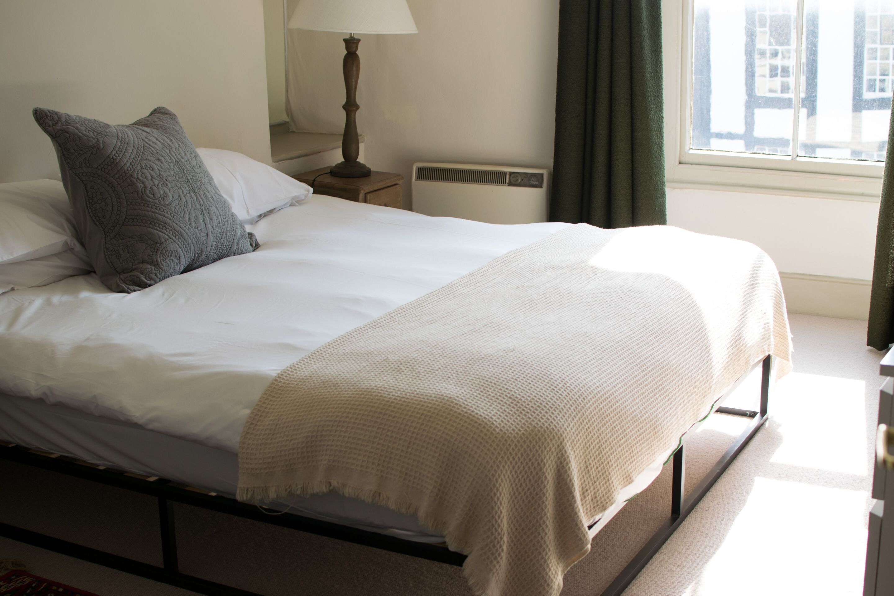 A neatly made bed with a white duvet and a grey pillow, a bedside lamp, a radiator, and a window with curtains.
