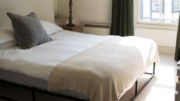A neatly made bed with a white duvet and a grey pillow, a bedside lamp, a radiator, and a window with curtains.