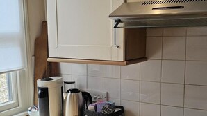 Une cuisine équipée d’une cuisinière, d’un four et de placards. On y trouve une fenêtre avec des stores, une bouilloire et un panier posé sur le comptoir.