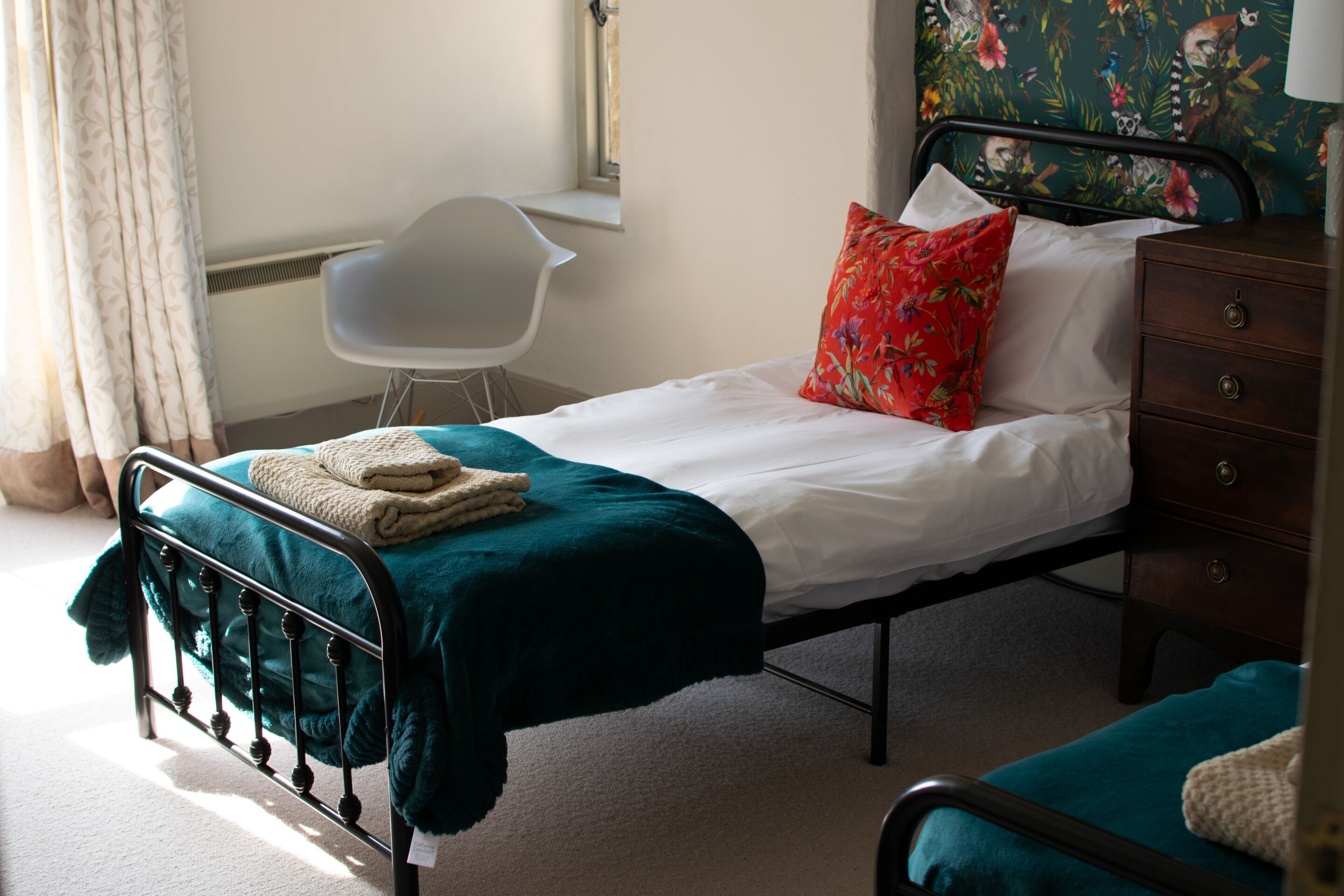 Una cama individual con cubrecama color turquesa, ropa de cama blanca y una almohada roja estampada. Se observa una silla blanca y un aparador de madera con cajones.