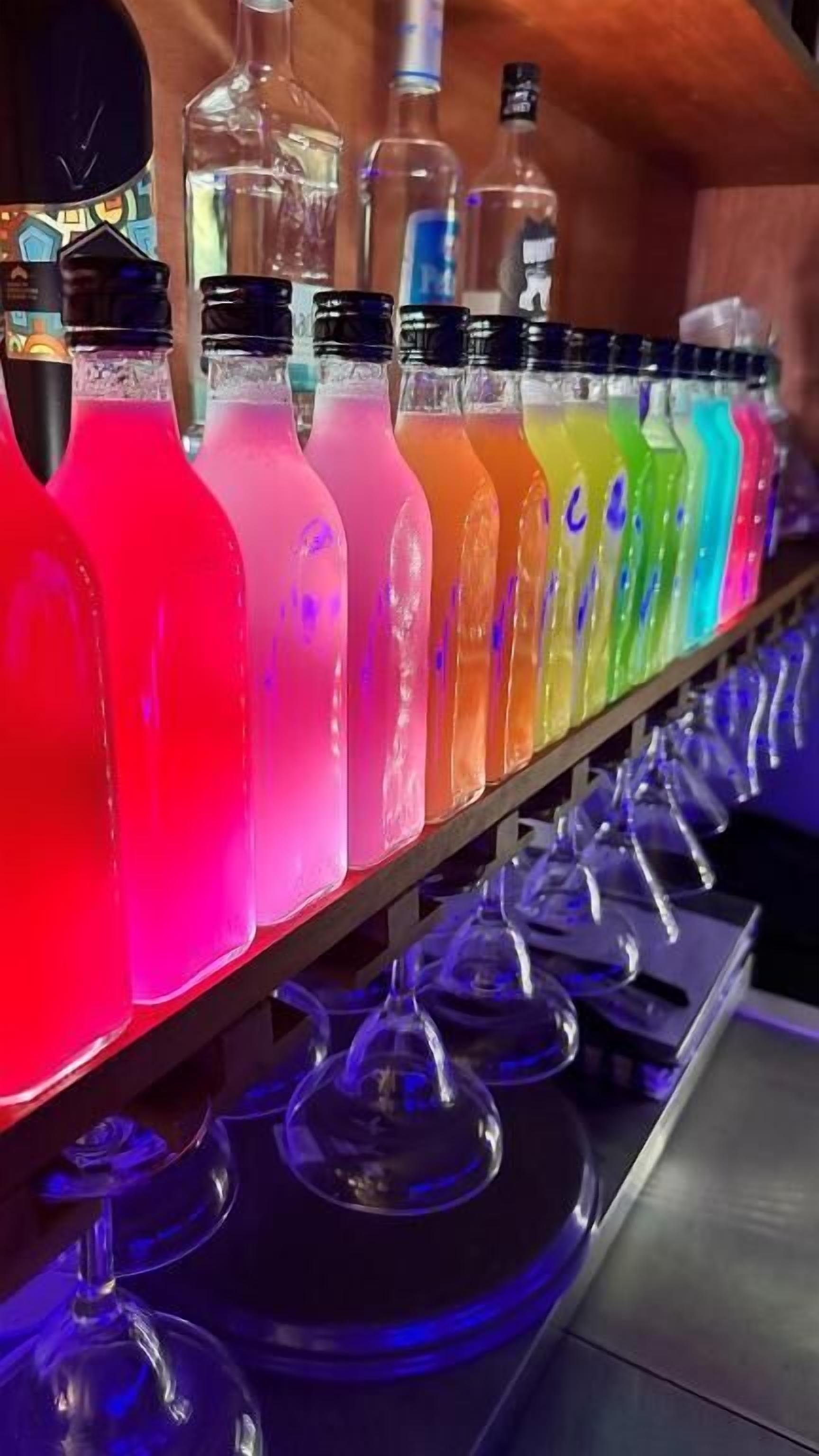 A row of colorful beverages on a shelf with glasses below.