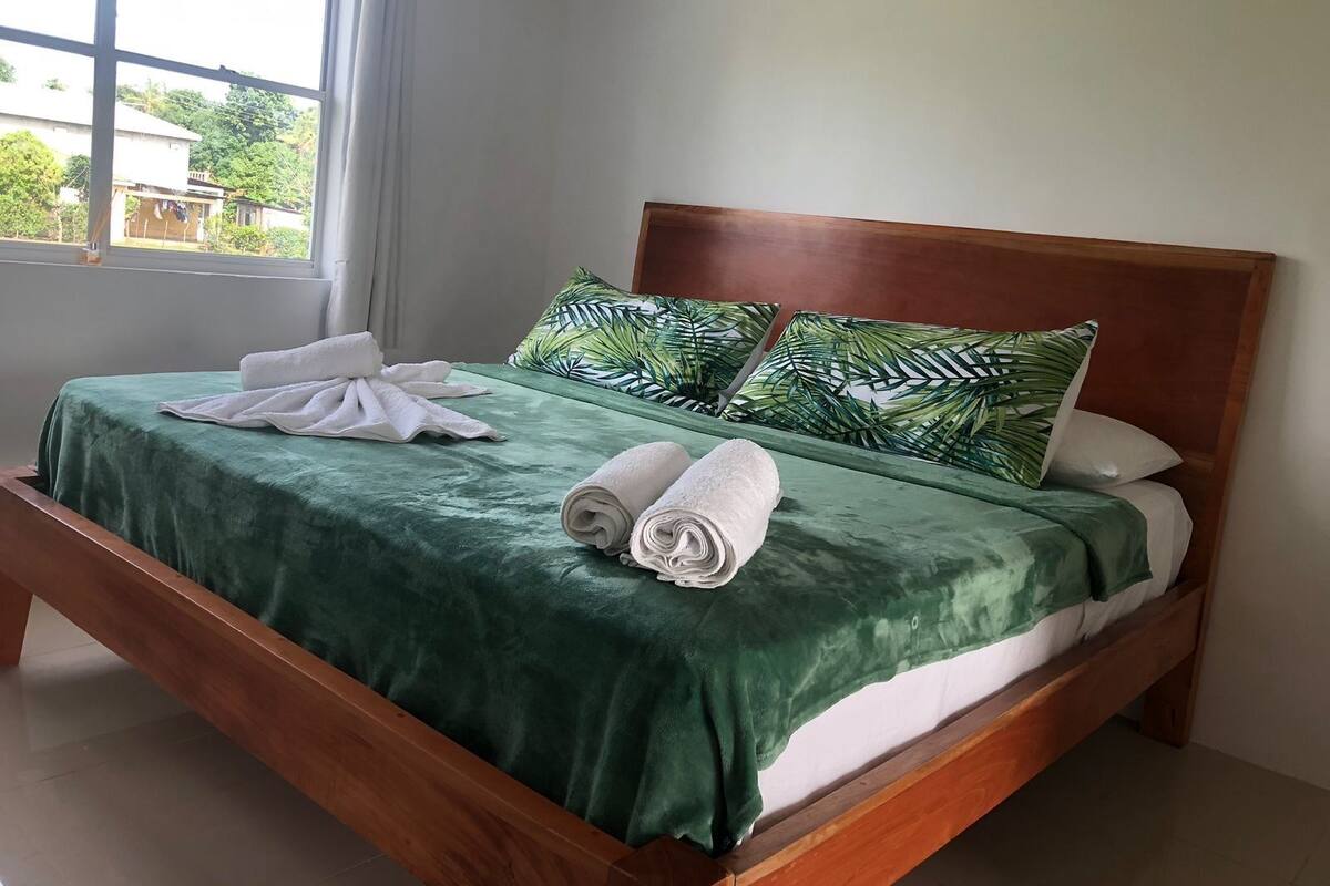 Una cama bien hecha con una colcha verde, dos toallas enrolladas y dos almohadas con un estampado de hojas tropicales.