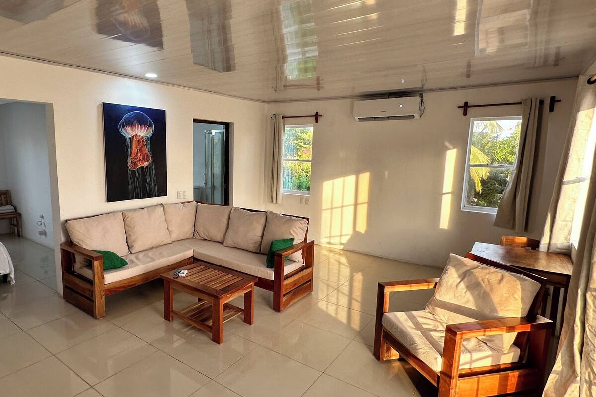 Amplia sala de estar con un sofá modular grande, una mesa de centro de madera y un sillón de madera. Hay un cuadro en la pared, una ventana con cortinas y una puerta que da a otra habitación.