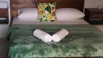 Una cama bien tendida con una colcha verde, dos almohadas blancas y una almohada con diseño de aguacate.