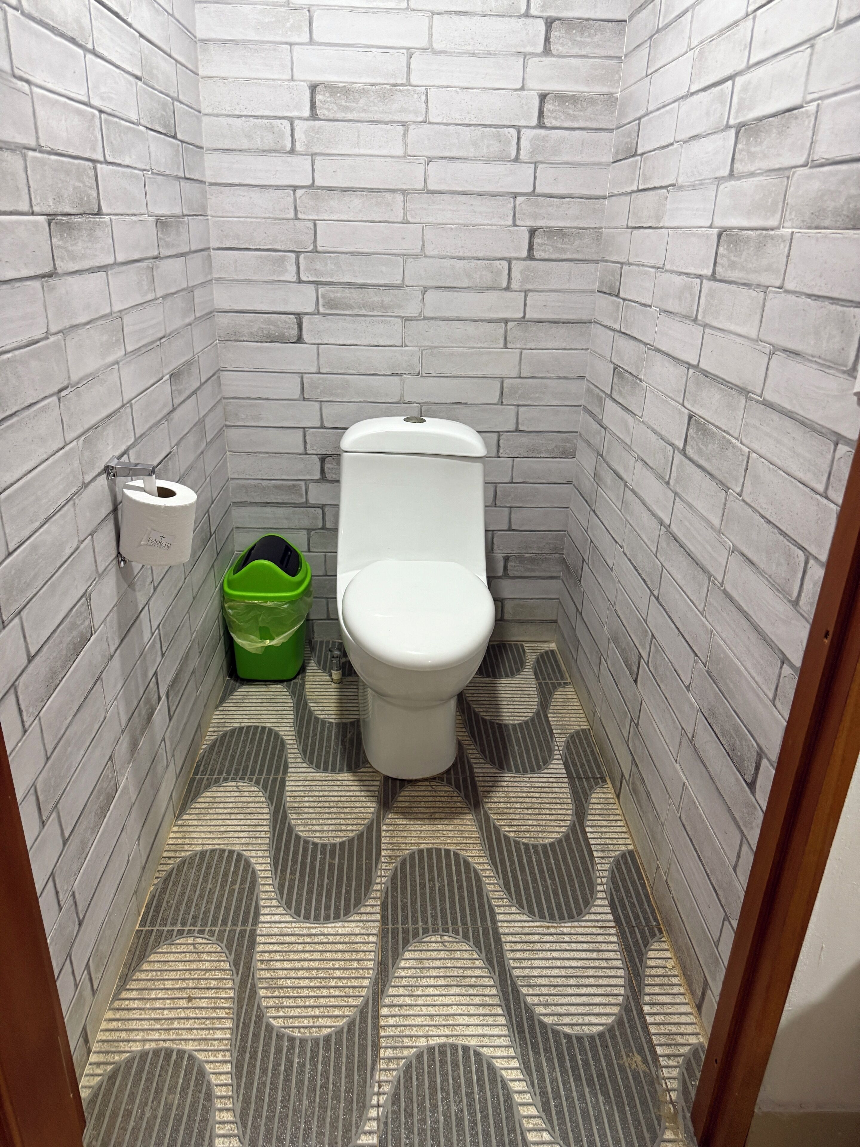Um banheiro pequeno e estreito, com um vaso sanitário branco, uma lixeira verde e um rolo de papel higiênico na parede.