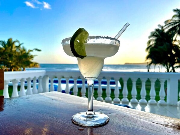 A margarita on a wooden table with a white balustrade and palm trees in the background. - Emerald Luxe Suites (Corn Islands)