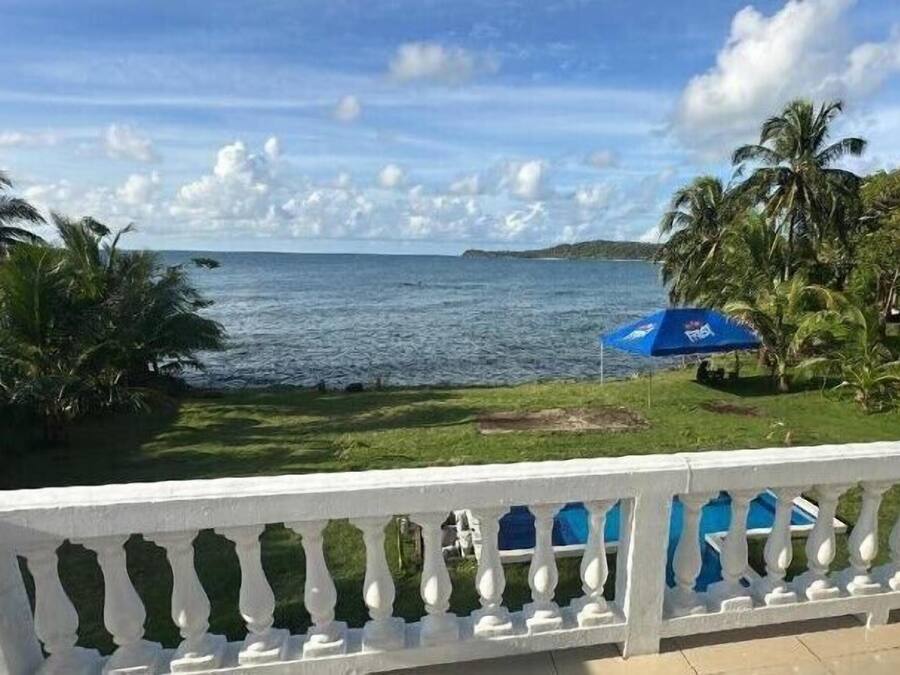 A balcony with a white balustrade overlooking a beach with palm trees and a blue umbrella.