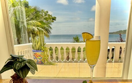 A breakfast plate with toast, bacon, eggs, and fruit, a glass of drink, and a view of the sea through a window.