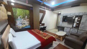 A hotel room with a bed, a sofa, a small table, a TV, and a kitchen area. - Shri Sai Palace (Mau Ranipur)