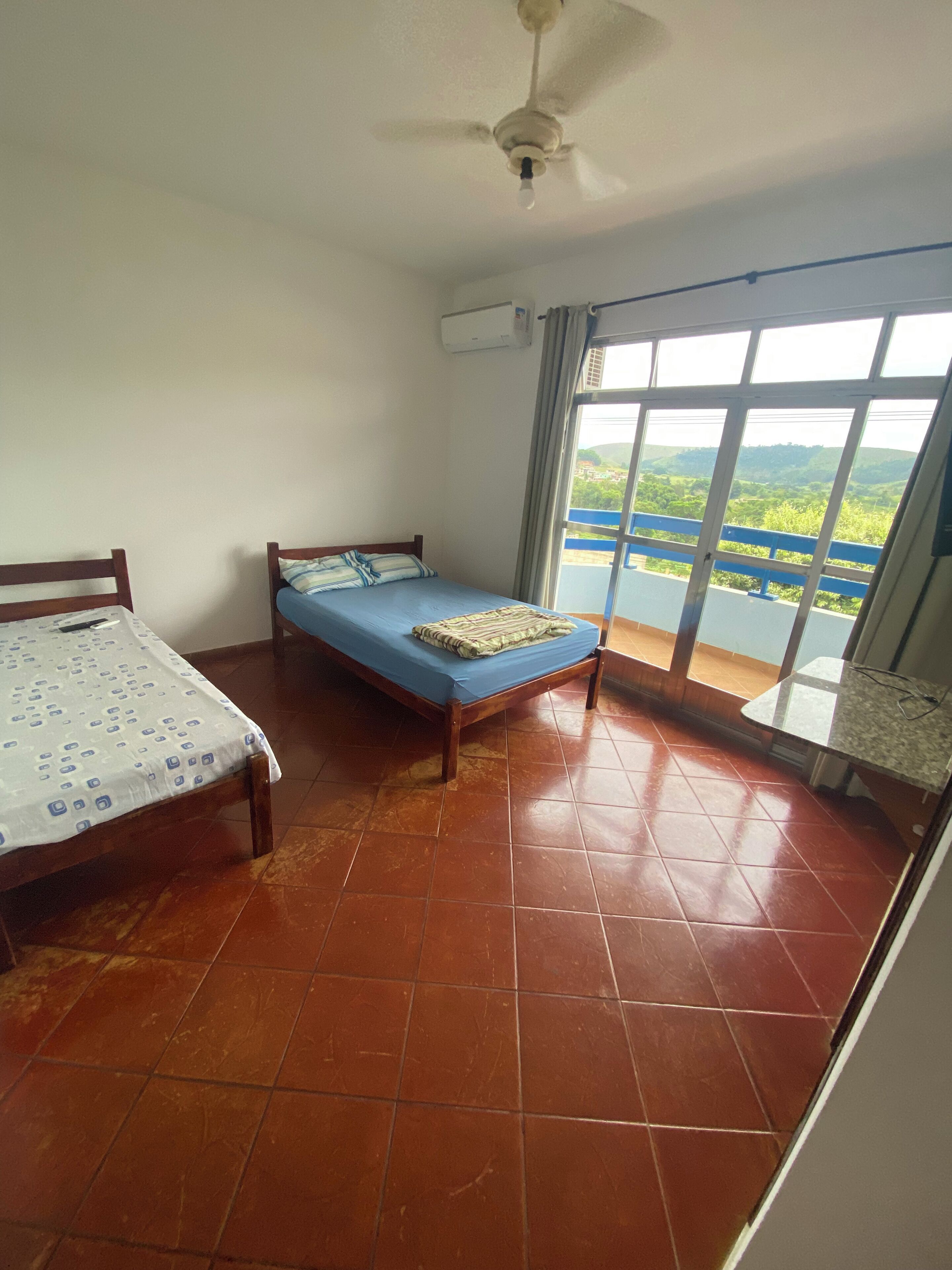 A room with two beds, a ceiling fan, and a large window with a view of greenery and hills.