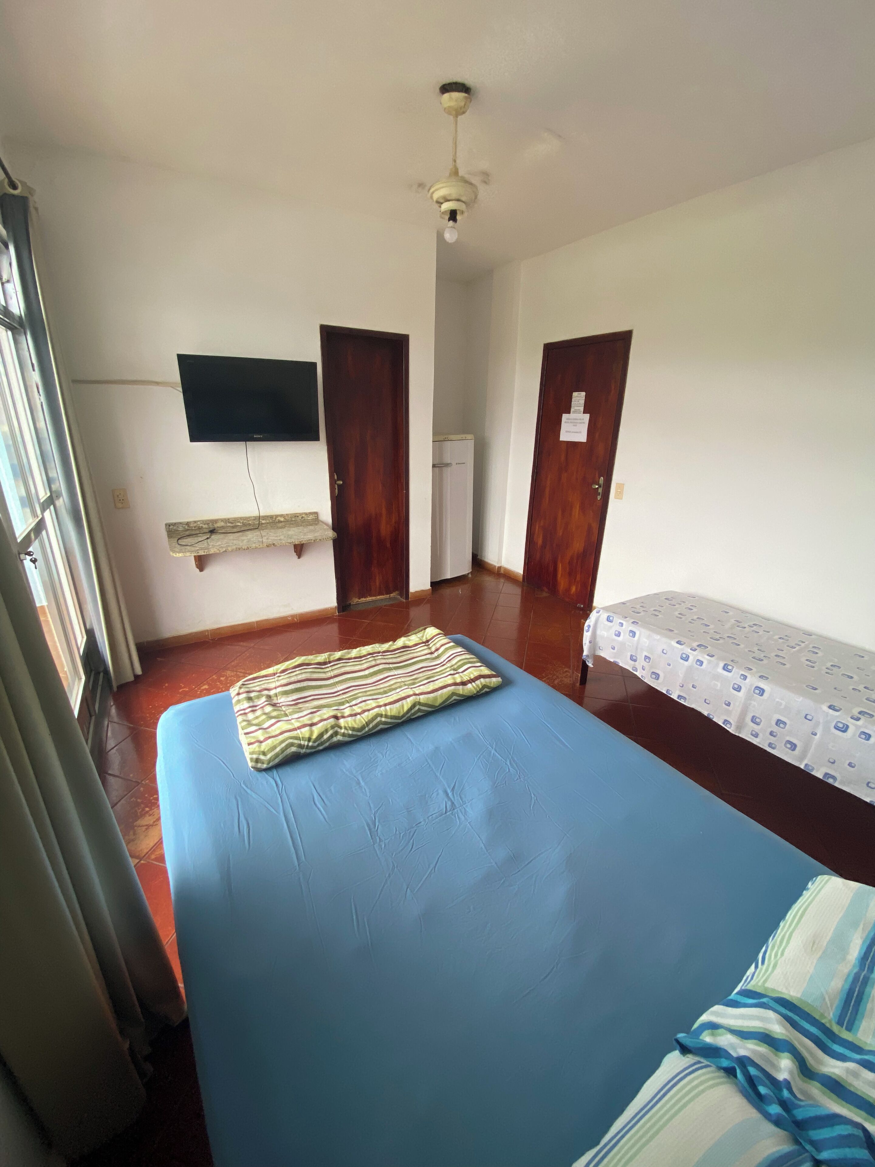 A room with a bed, a TV mounted on the wall, a ceiling fan, and a window.