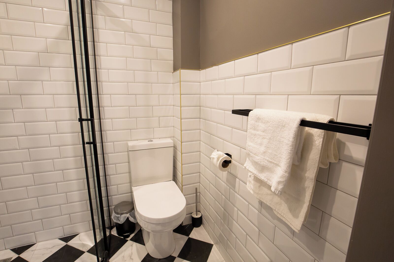 Ein Badezimmer mit weißen Fliesen im U-Bahn-Stil, einem schwarz-weißen Schachbrettboden, einer weißen Toilette, einem Handtuchhalter mit einem weißen Handtuch und einem Mülleimer.