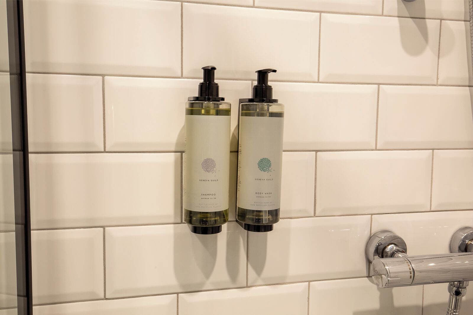 Dos botellas de champú y gel de baño Geneva Guild sobre una pared de azulejos blancos.