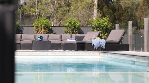 A poolside area with a dark-colored sofa set and lounge chairs, a small table with a bottle and glasses, and a blue towel.
